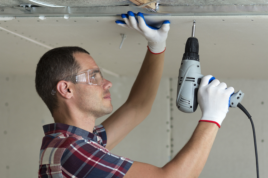 Young Man In Goggles Fixing Drywall Suspended Ceiling To Metal F Young man in goggles fixing drywall suspended ceiling to metal frame using electrical screwdriver on ceiling insulated with shiny aluminum foil. Renovation, construction, do it yourself concept.
