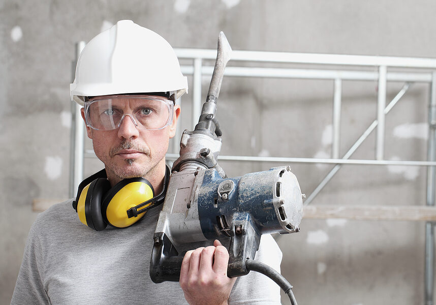 portrait of man construction worker with jackhammer with safety hard hat, hearing protection headphones and protective glasses. look at the camera isolated on interior building site background
