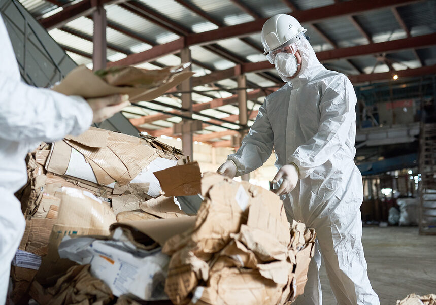 Portrait of  factory worker wearing biohazard suit sorting recyclable cardboard on waste processing plant, copy space